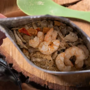 A close-up view of a hand holding an opened bag of freeze-dried food containing shrimp and assorted vegetables on a wooden surface. A green fork is placed beside the bag.