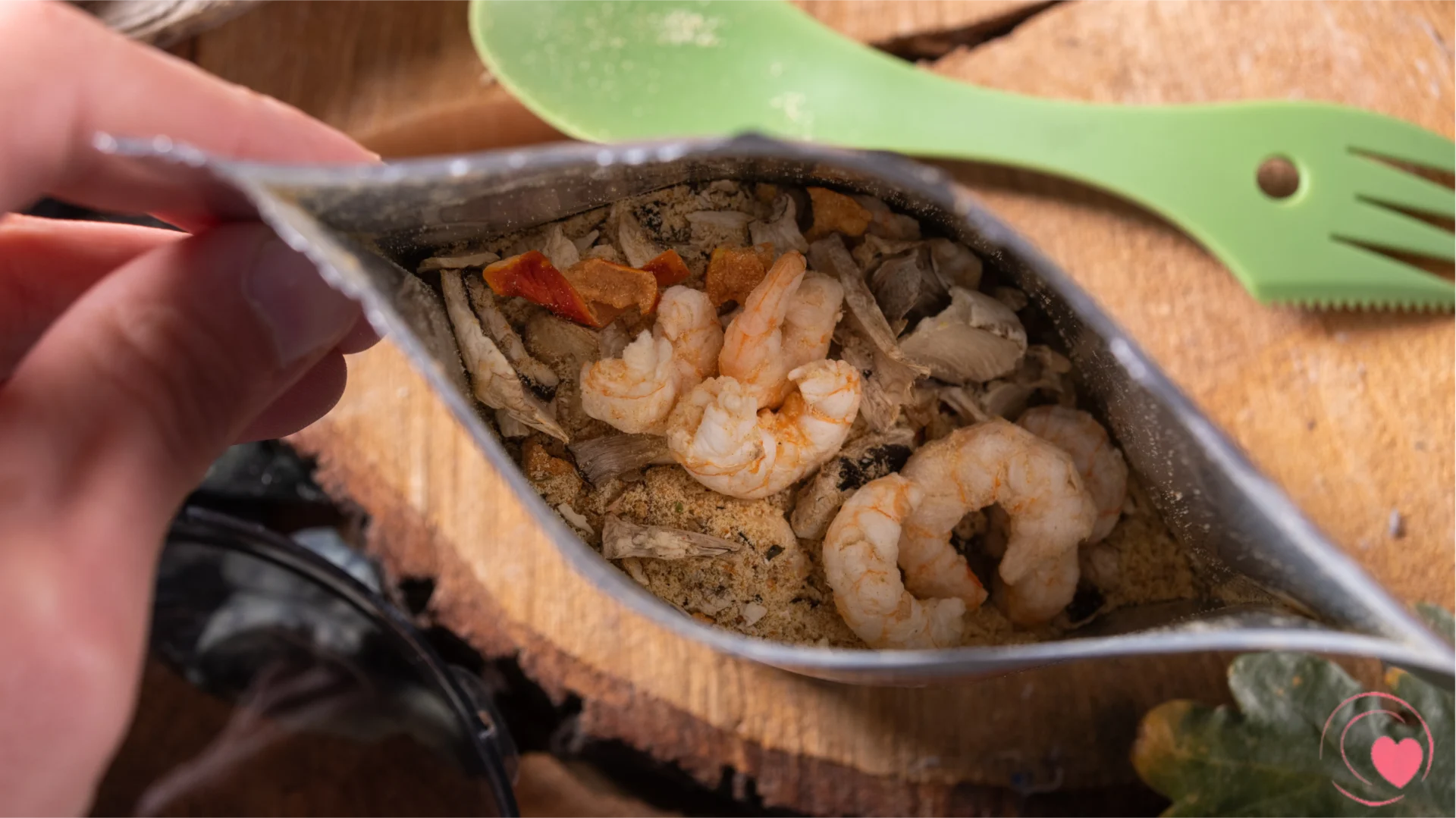 A close-up view of a hand holding an opened bag of freeze-dried food containing shrimp and assorted vegetables on a wooden surface. A green fork is placed beside the bag.